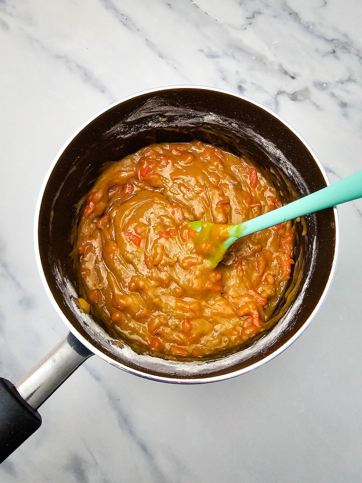 Overhead view of a dark cooking pot filled with a thick caramel-colored mixture speckled with orange pieces, likely shredded carrots, being stirred with a light blue silicone spatula, on a white marble surface.