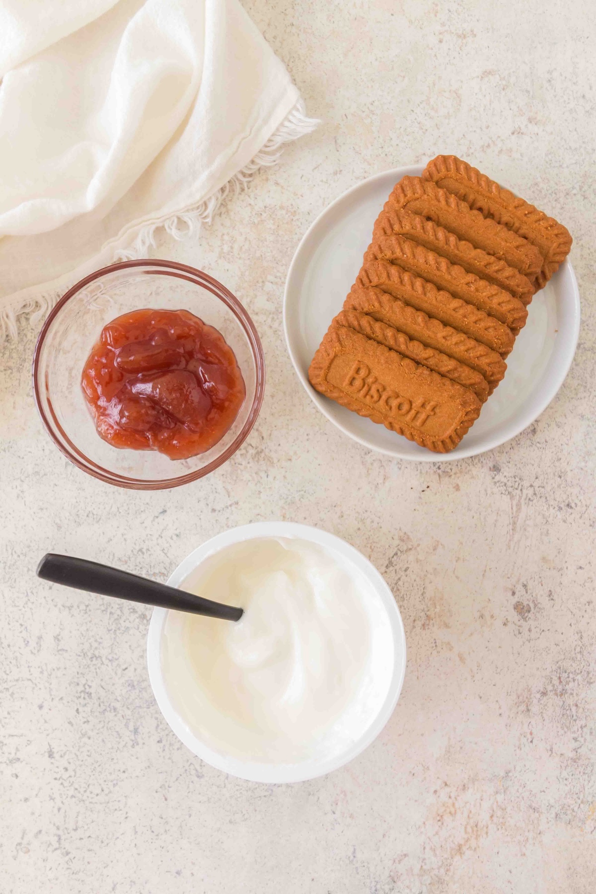 Overhead view of Biscoff biscuits, jam, and yogurt in bowls on a light surface with a white cloth.