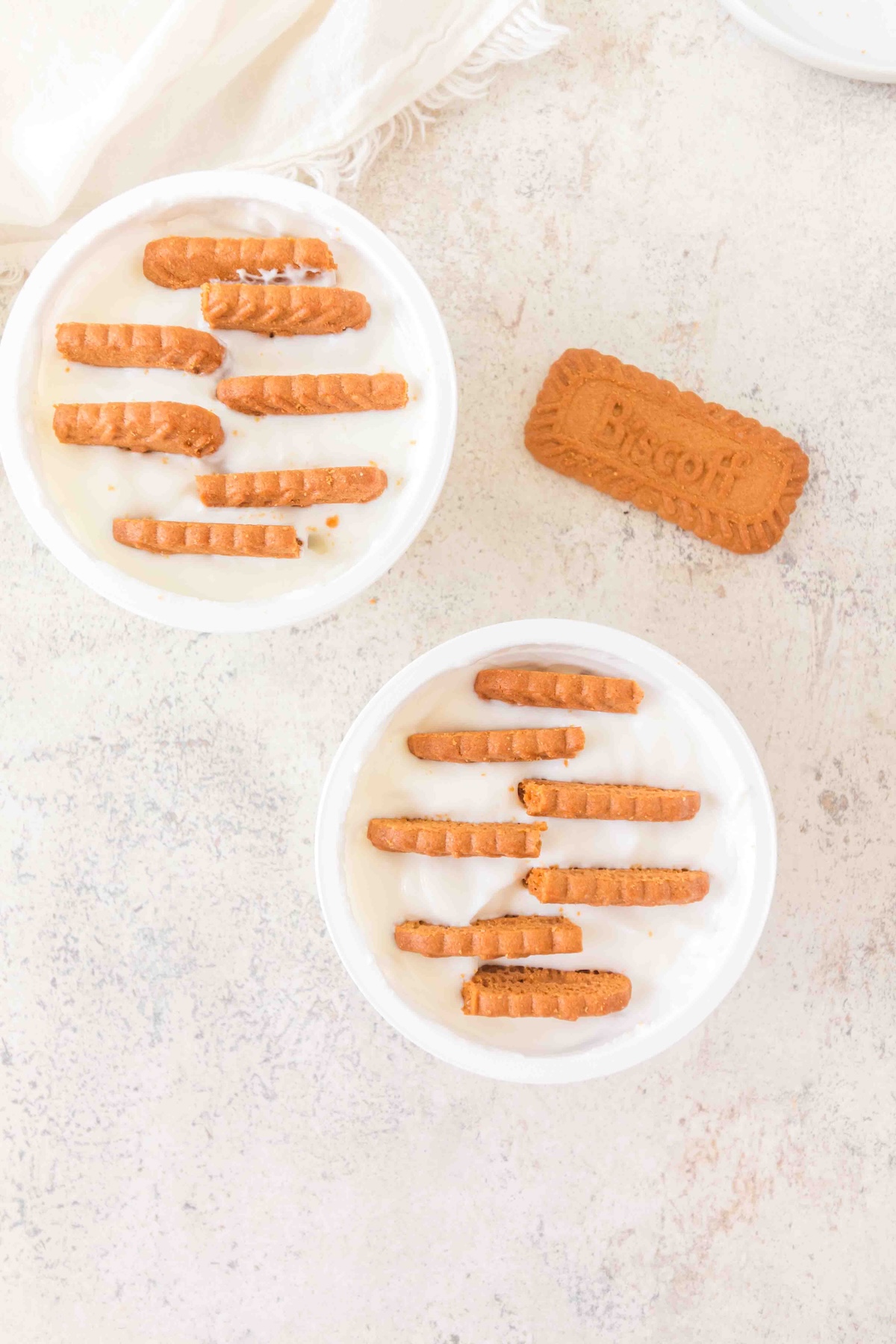 Two bowls of yogurt with Biscoff cookies, a whole cookie on a light surface, and a white cloth in the corner.