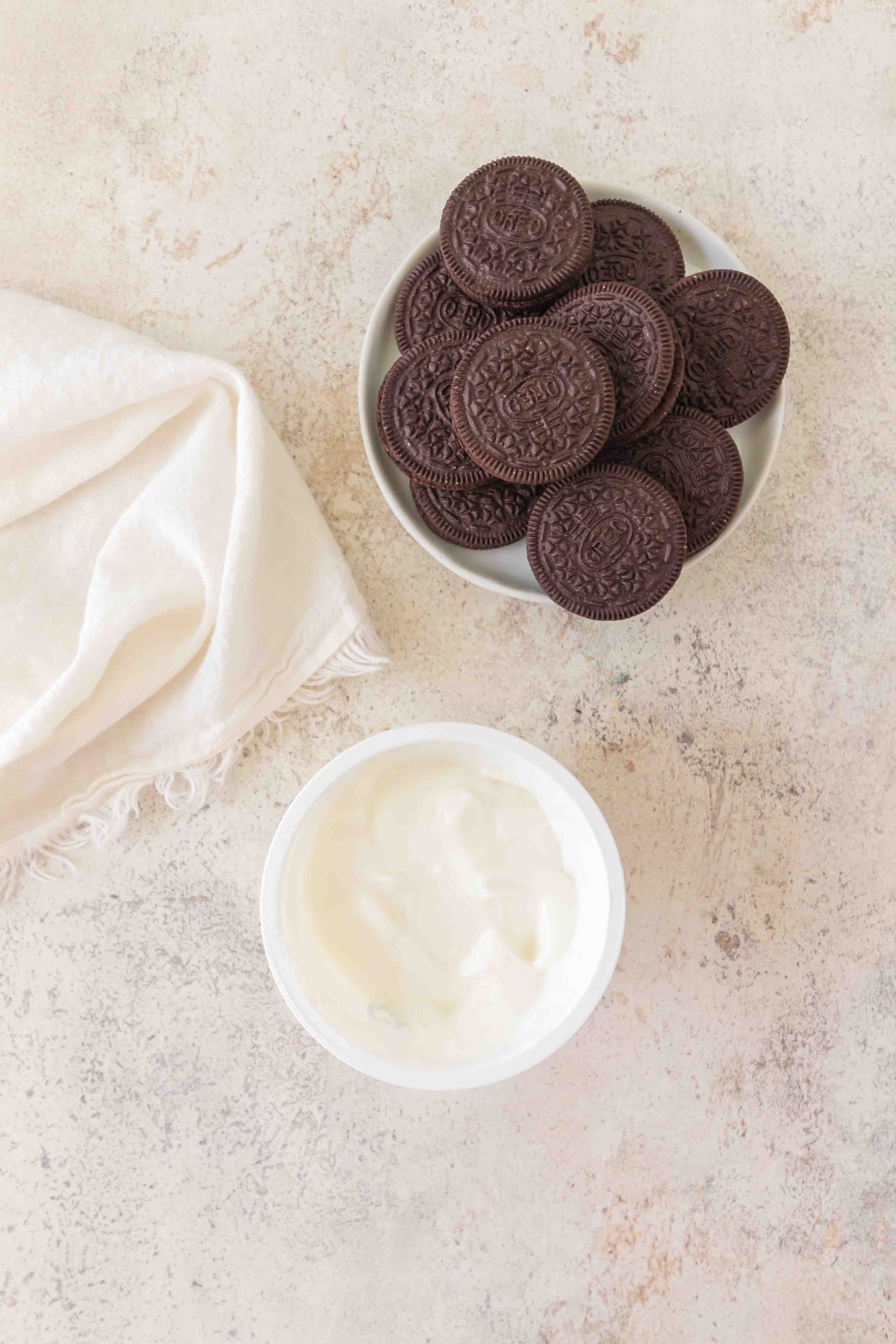 Plate of Oreo cookies, a bowl of yogurt, and a white cloth on a textured light surface.