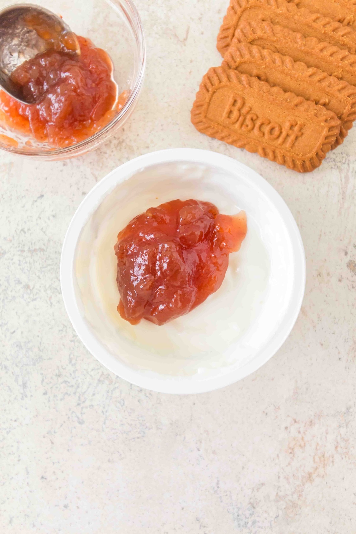 Overhead view of a white bowl with yogurt topped with reddish-orange fruit preserve, a glass bowl of preserve with a spoon, and a stack of three Biscoff biscuits on a light textured surface.