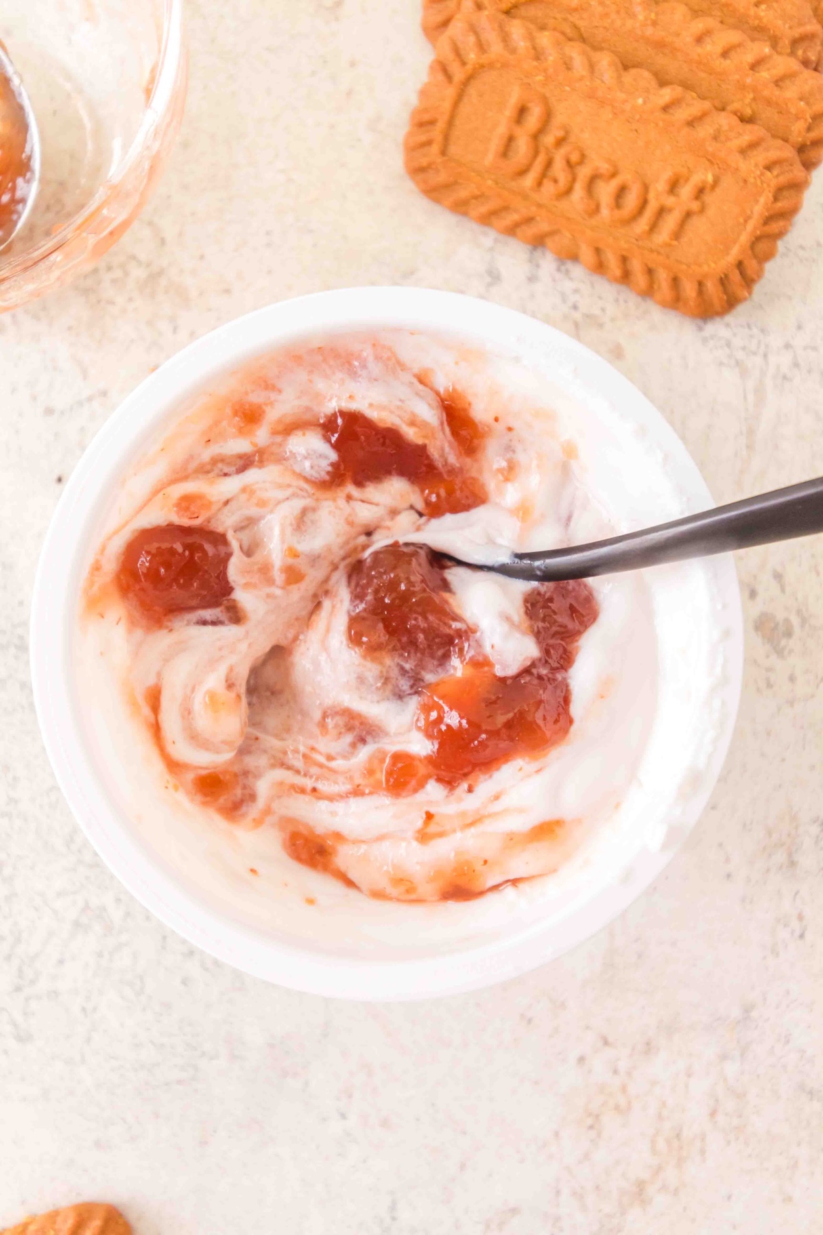 Overhead view of a white bowl with yogurt swirled with reddish-orange jam and a dark spoon, a small glass bowl of jam in the corner, and three stacked Biscoff biscuits on a light textured surface.