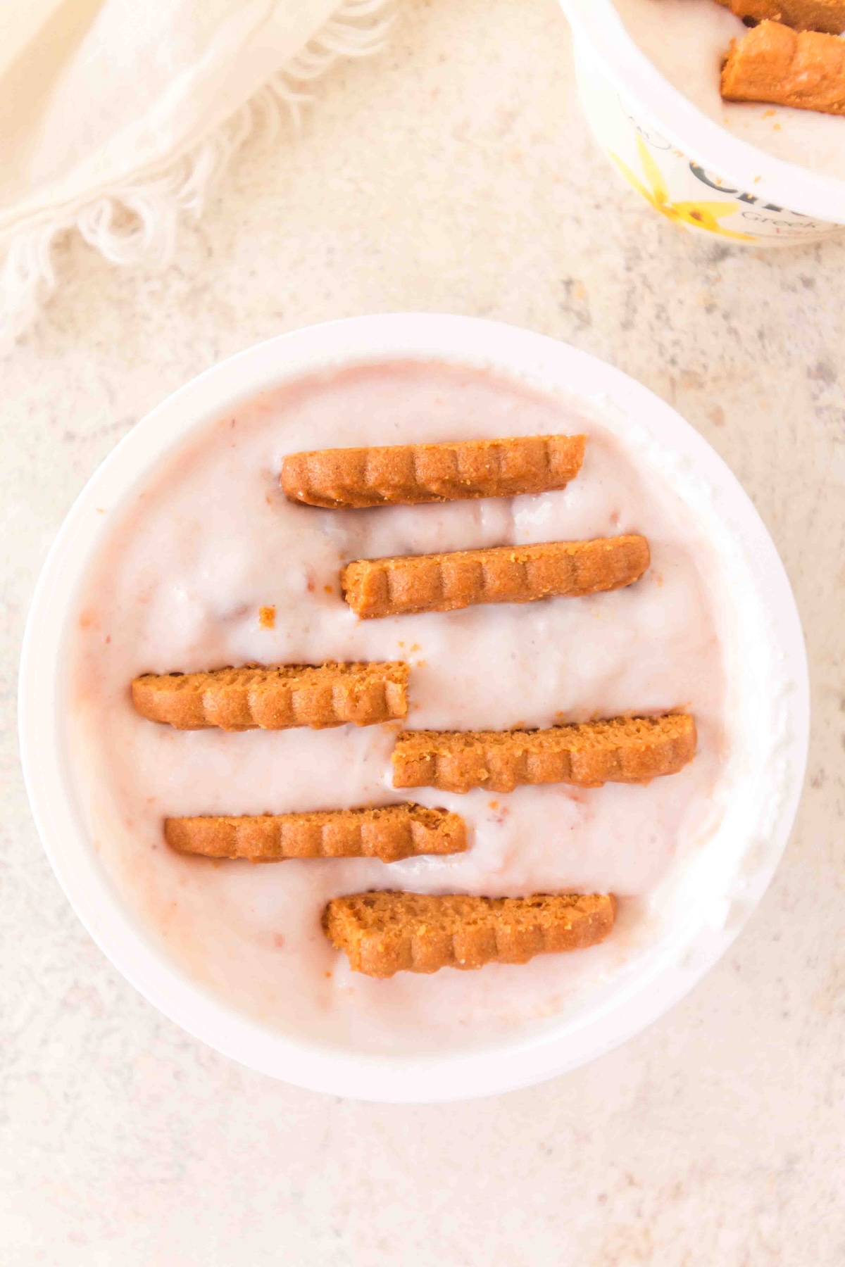 Overhead view of a white bowl filled with light pink yogurt and seven golden-brown biscuits partially submerged, on a light textured surface with a white fringed cloth in the corner. Another bowl with biscuits and a floral rim is partially visible in the background.
