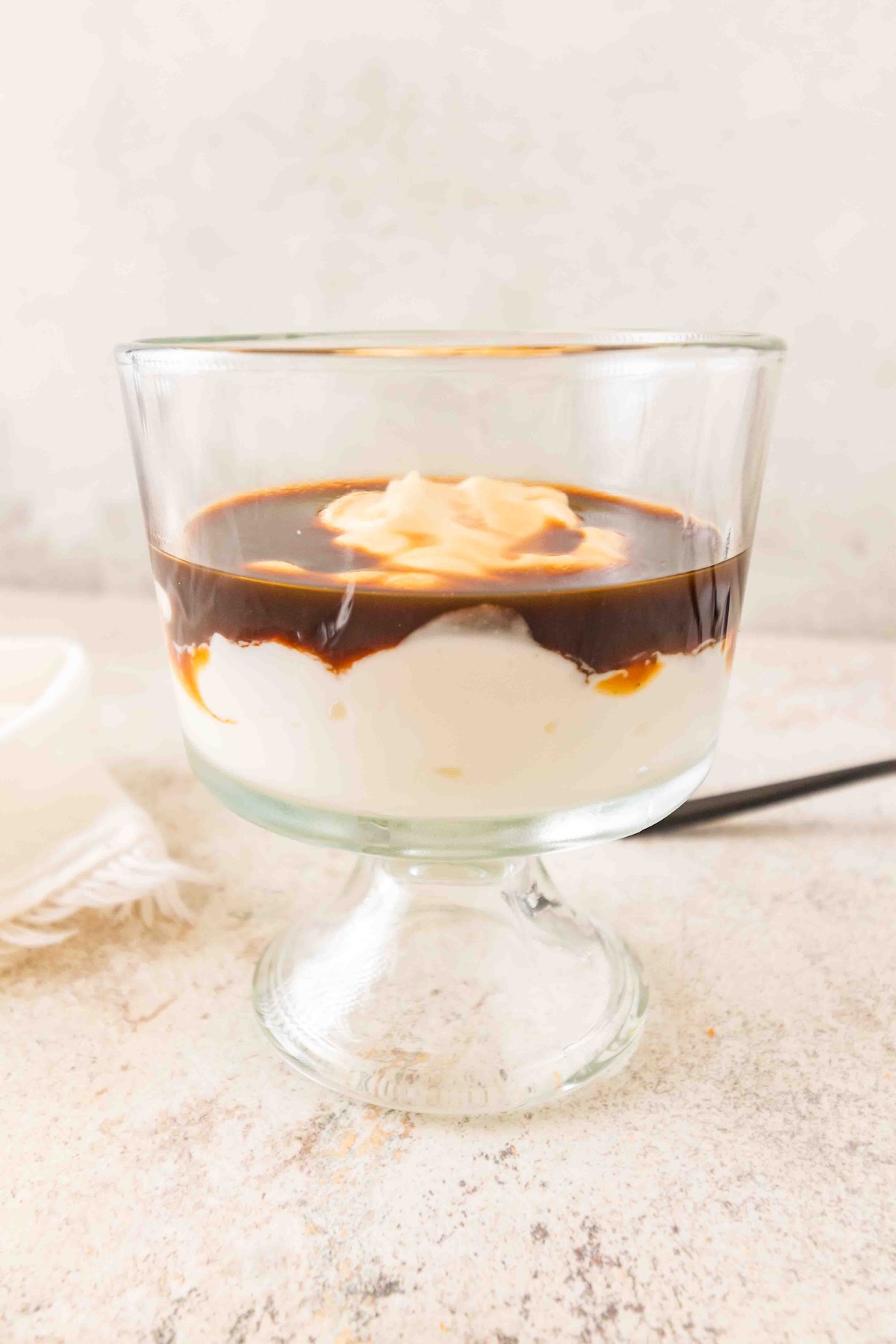 Layered dessert in a clear glass cup with a creamy base, dark sauce, and dollops of cream, set on a light surface with a napkin nearby.