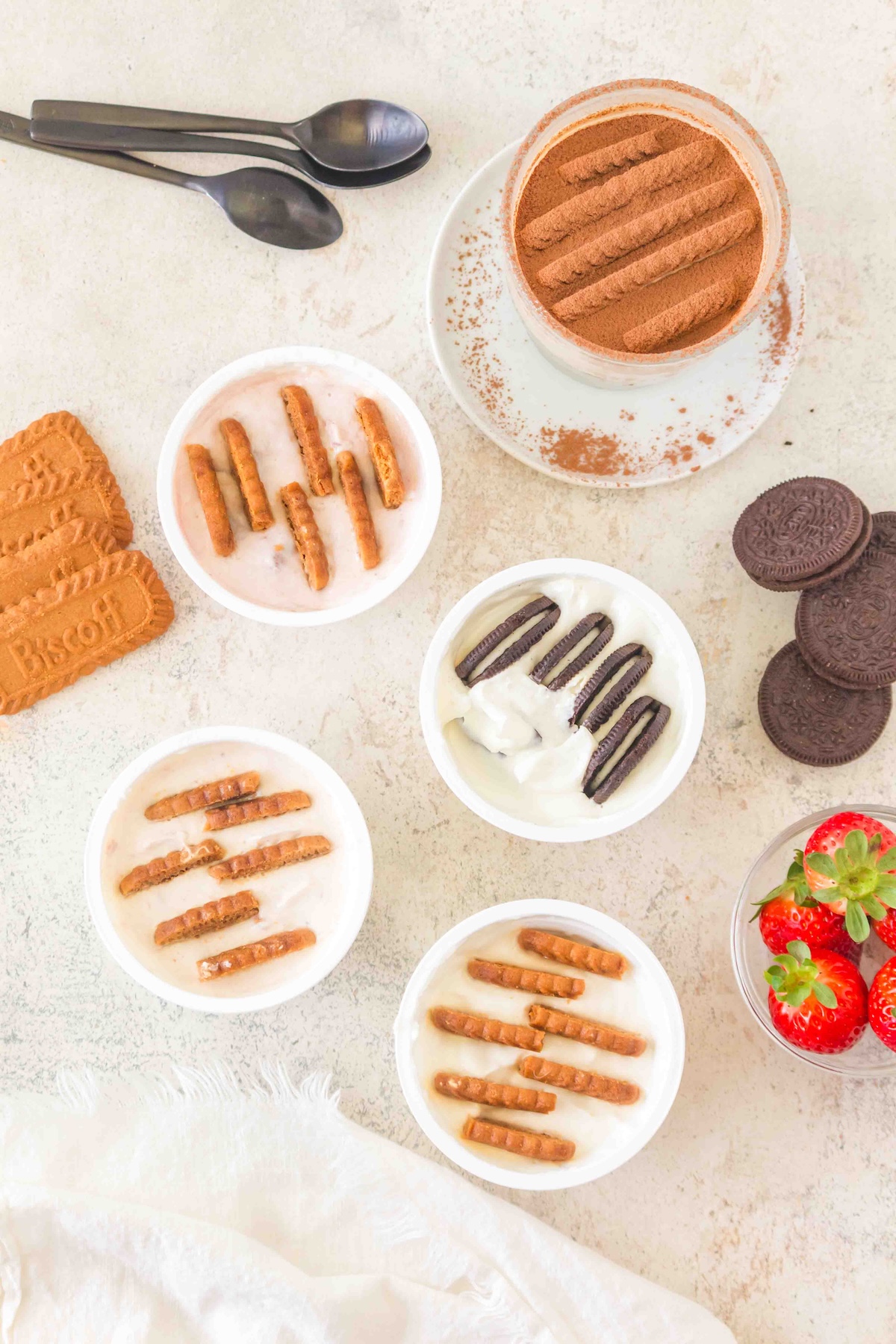 Flat lay of desserts in bowls topped with biscuits or cookies, a stack of Biscoff cookies, and fresh strawberries on a light surface.