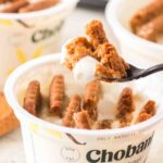 Chobani Vanilla Greek Yogurt cup with biscuit pieces, a spoon lifting a portion, and additional yogurt cups in the background.