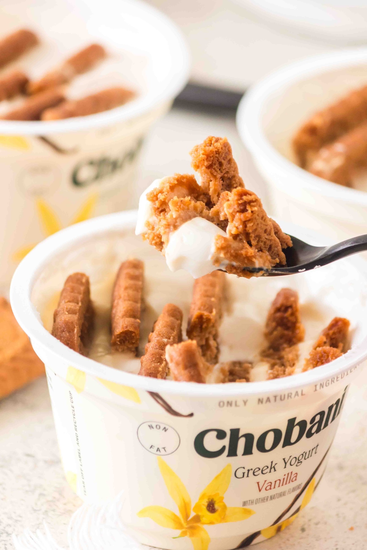 Chobani Vanilla Greek Yogurt cup with biscuit pieces, a spoon lifting a portion, and additional yogurt cups in the background.