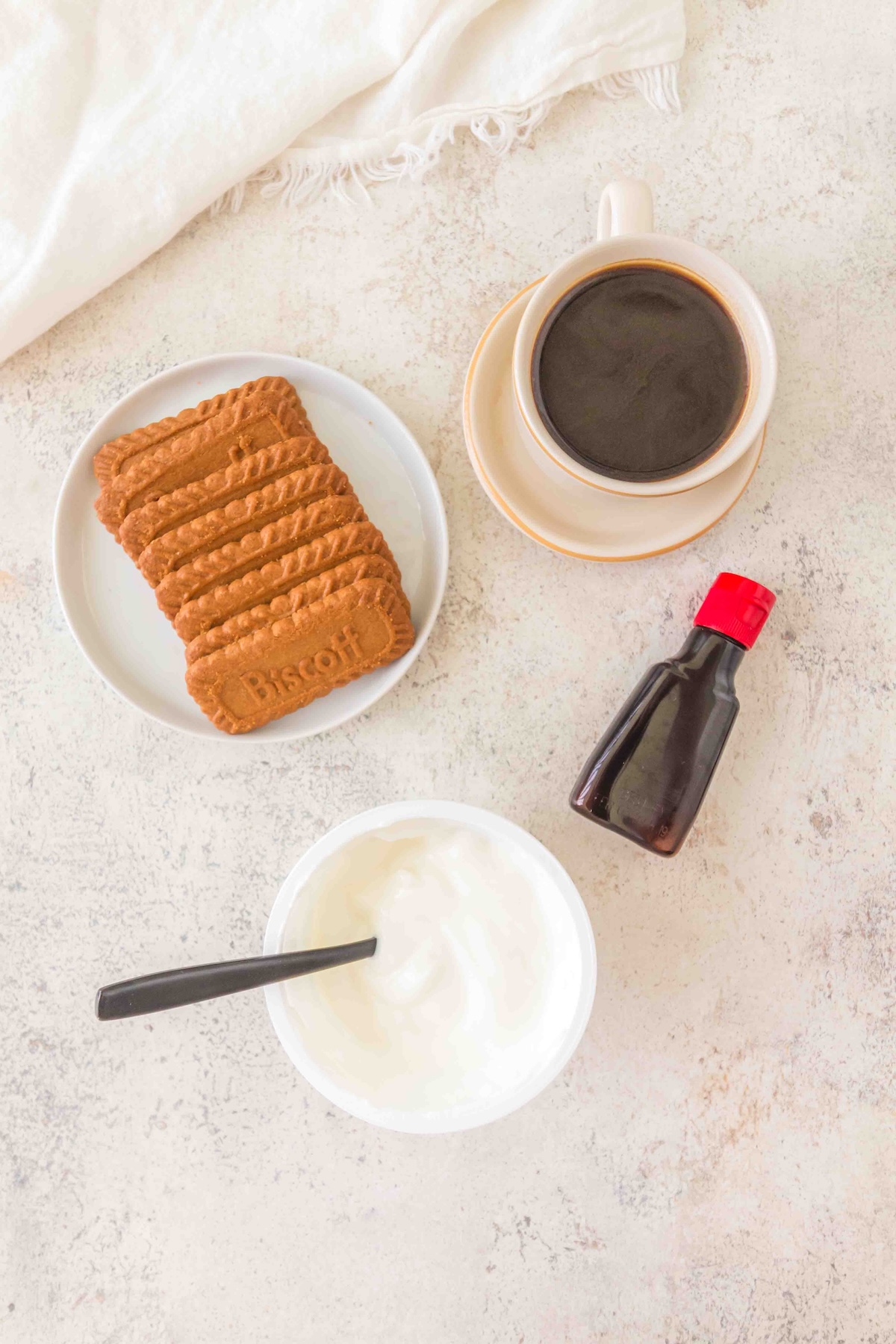 Close-up flat lay: stack of Biscoff cookies on plate, Chobani vanilla yogurt tub, coffee cup, vanilla extract bottle, cozy white fabric backdrop.