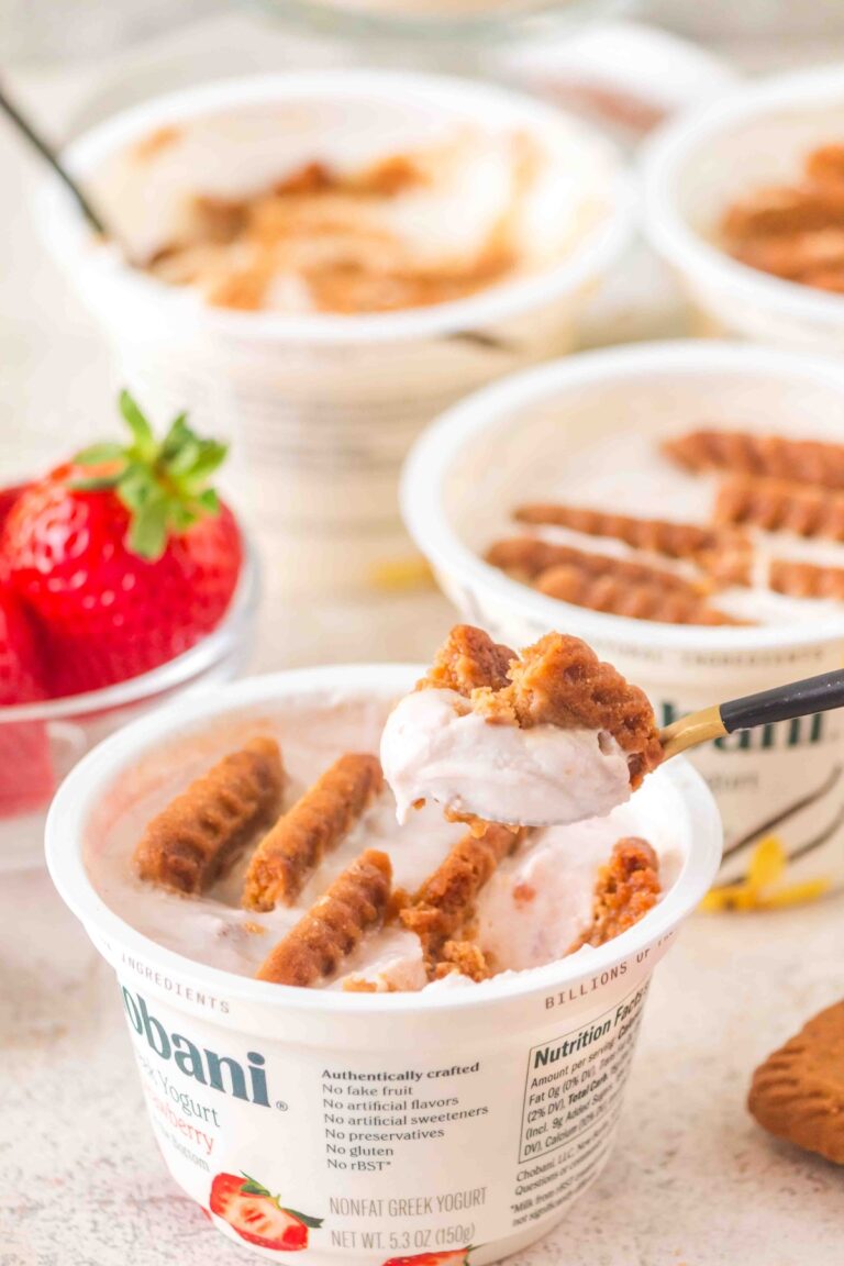 Spoon lifting strawberry yogurt with crunchy biscuit pieces from a Chobani cup, fresh strawberries nearby.