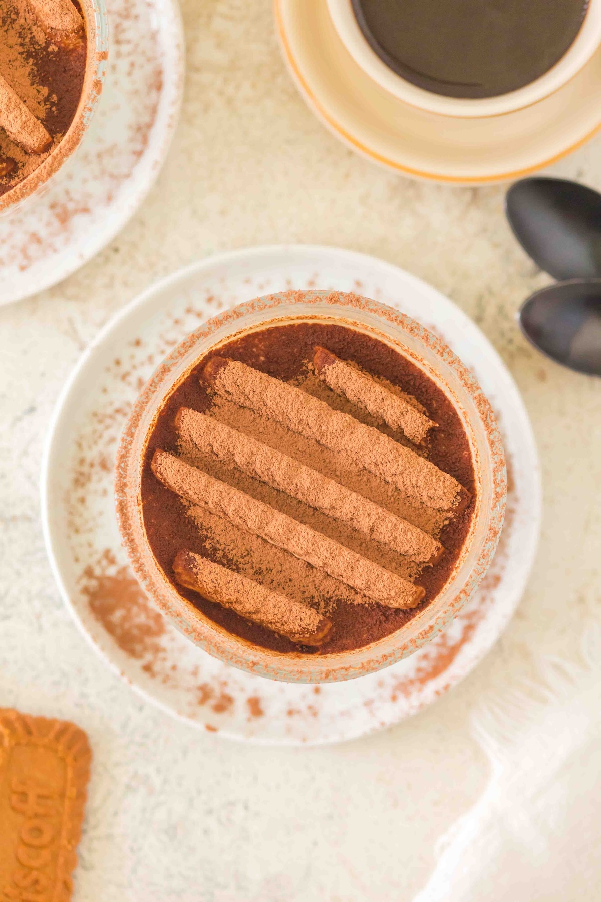 Tiramisu in a glass with cocoa-dusted biscuits, coffee cup, and a Biscoff cookie on a light plate.