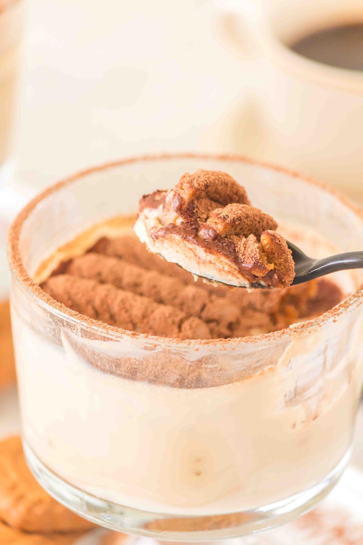 Glass cup of tiramisu with creamy layers, cocoa powder, and a spoon lifting a rich, textured scoop.
