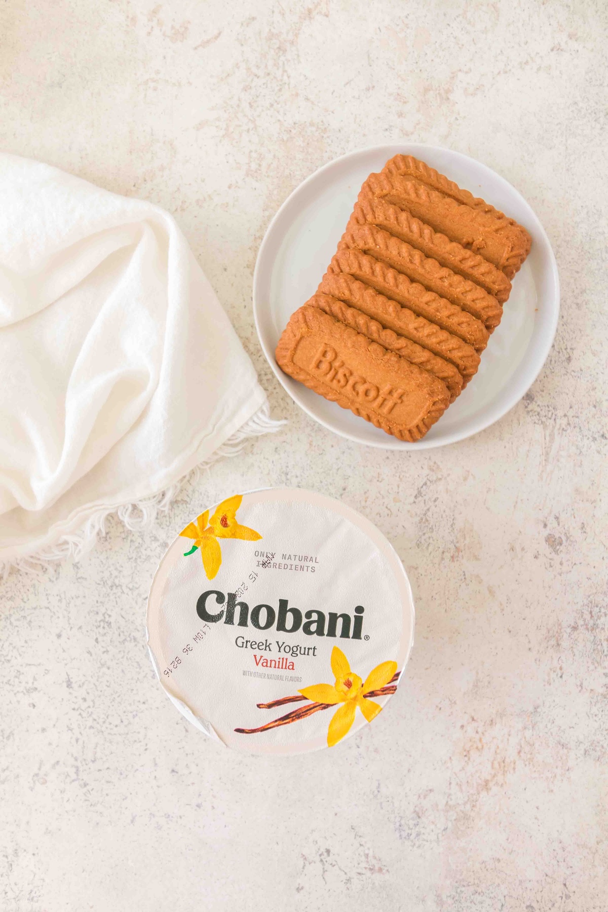 Chobani vanilla Greek yogurt tub topped with stacked Biscoff cookies on white plate, soft white fabric and orchid accents.