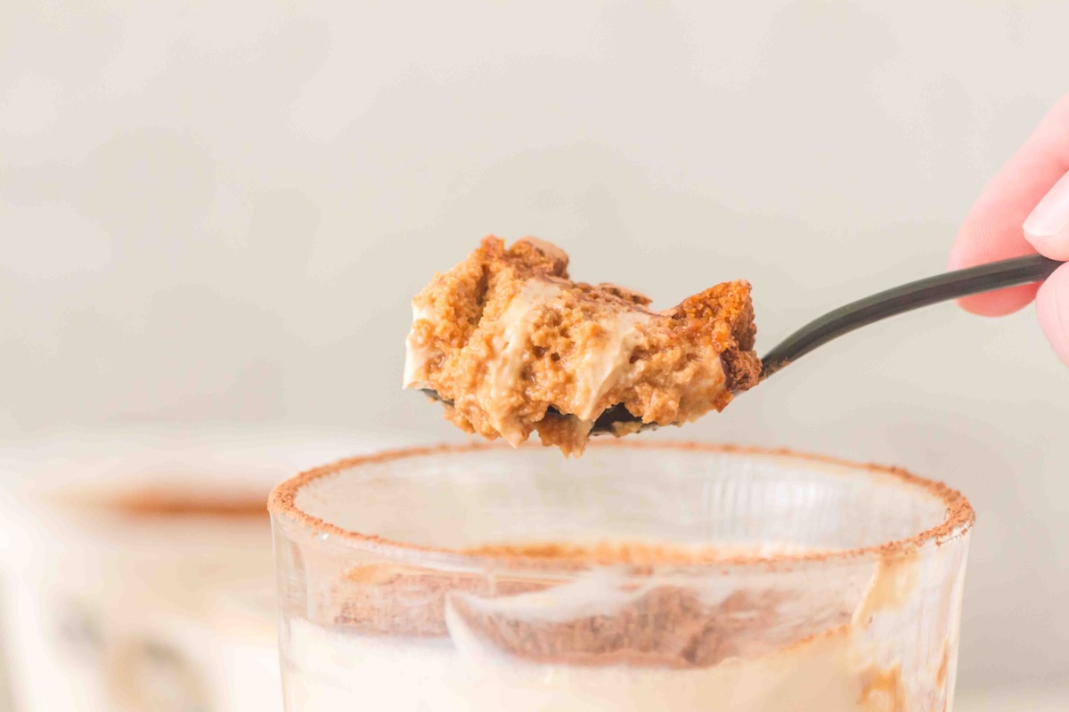 Spoon holding a layered dessert above a glass, showcasing creamy and crumbly textures with a cocoa-dusted rim.
