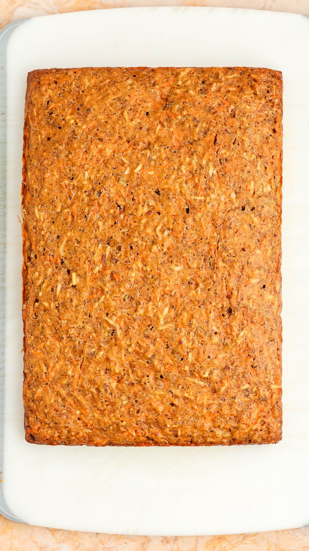 Overhead view of a rectangular carrot cake loaf with a textured orange-brown surface, resting on a white cutting board placed on a light marble countertop. The cake shows visible shreds of carrots and specks of nuts or spices.