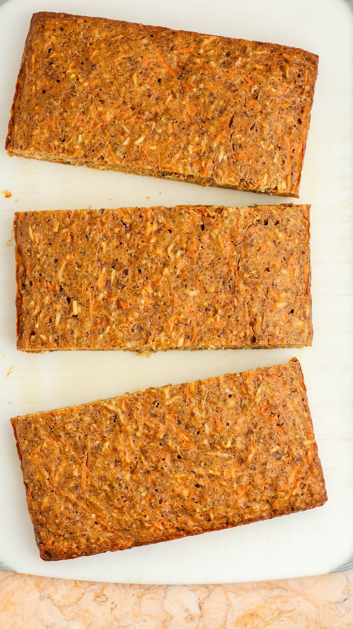Top-down shot of three rectangular carrot cake loaves with textured orange-brown surfaces, arranged vertically on a white cutting board. The background features a light marble countertop with warm tones.