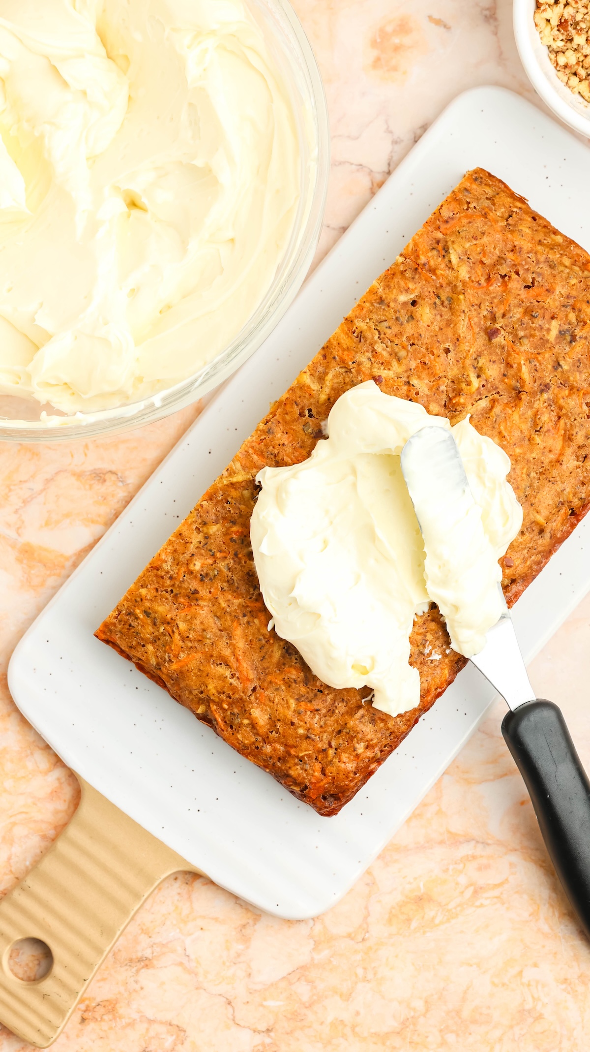 Overhead view of a rectangular carrot cake loaf being frosted with creamy white icing using a metal spatula. A bowl of frosting and a bowl of chopped nuts are visible in the background, all set on a marbled countertop.
