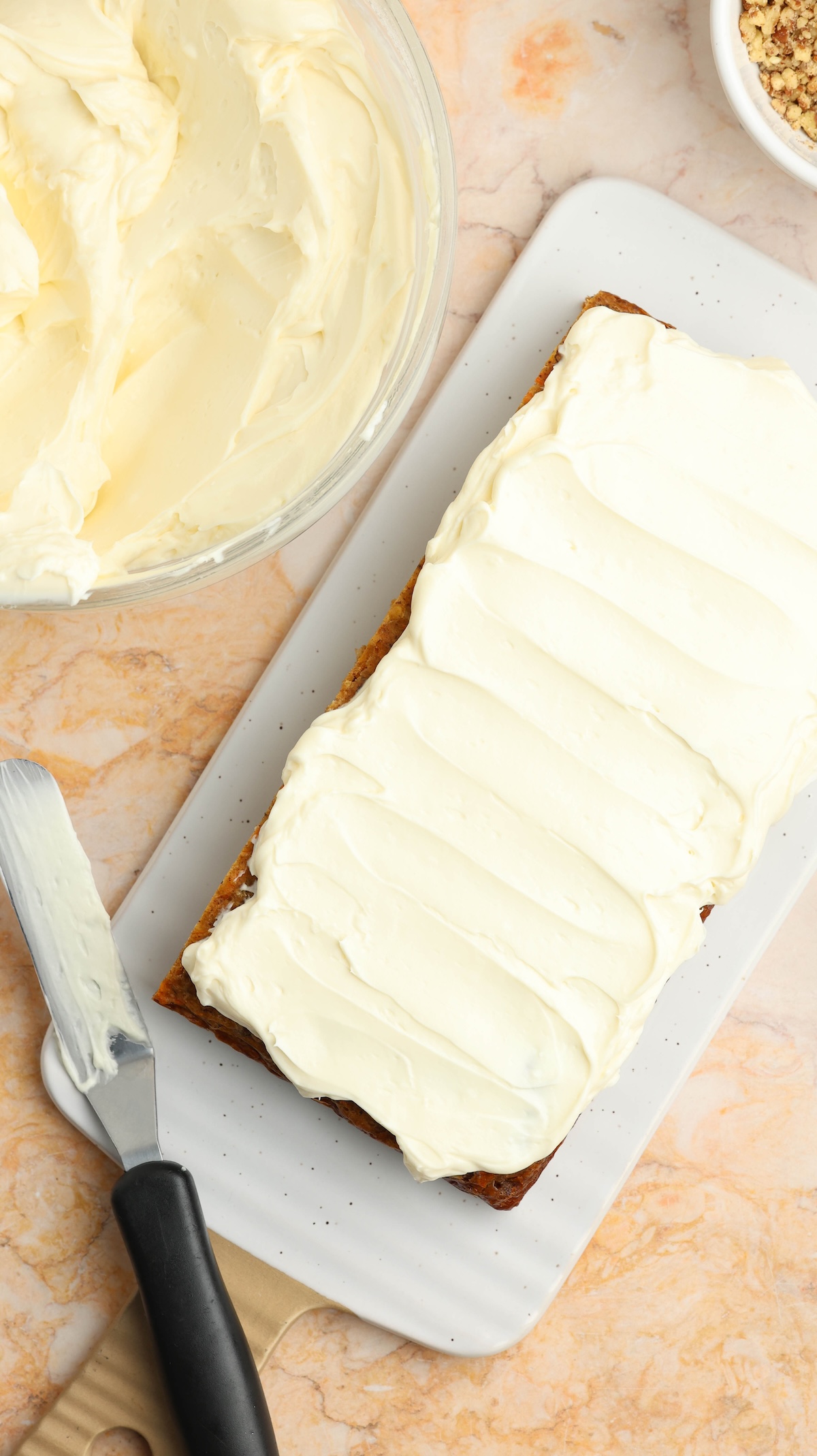 Flat lay of a frosted rectangular carrot cake on a white serving platter. A metal spatula with frosting rests nearby, alongside a glass bowl of frosting and a bowl of chopped nuts, all on a marbled countertop.