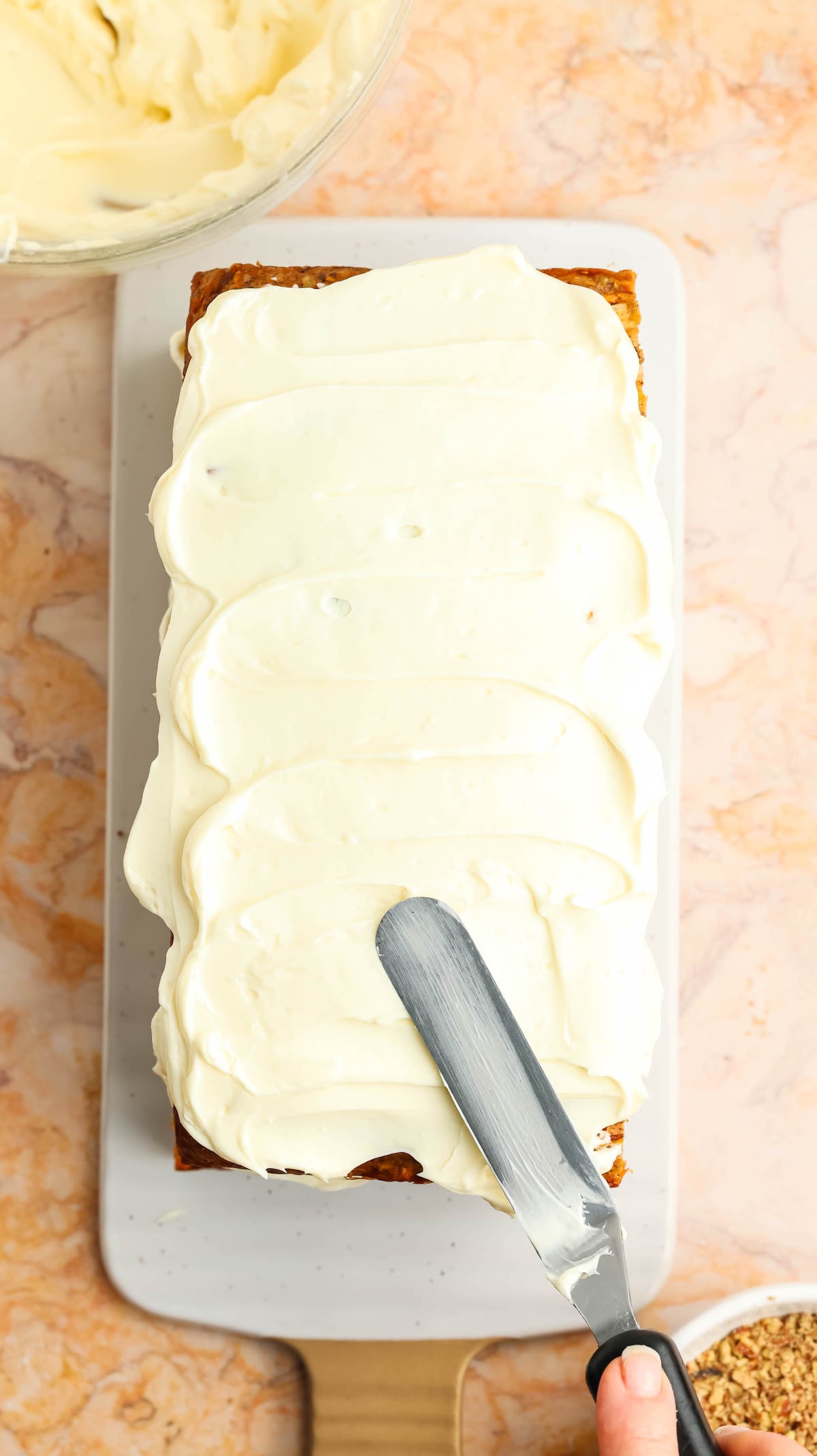 Overhead view of a rectangular carrot cake being frosted with creamy white icing. A hand holding a metal spatula is spreading the frosting, with a bowl of frosting and a bowl of chopped nuts nearby on a marbled countertop.