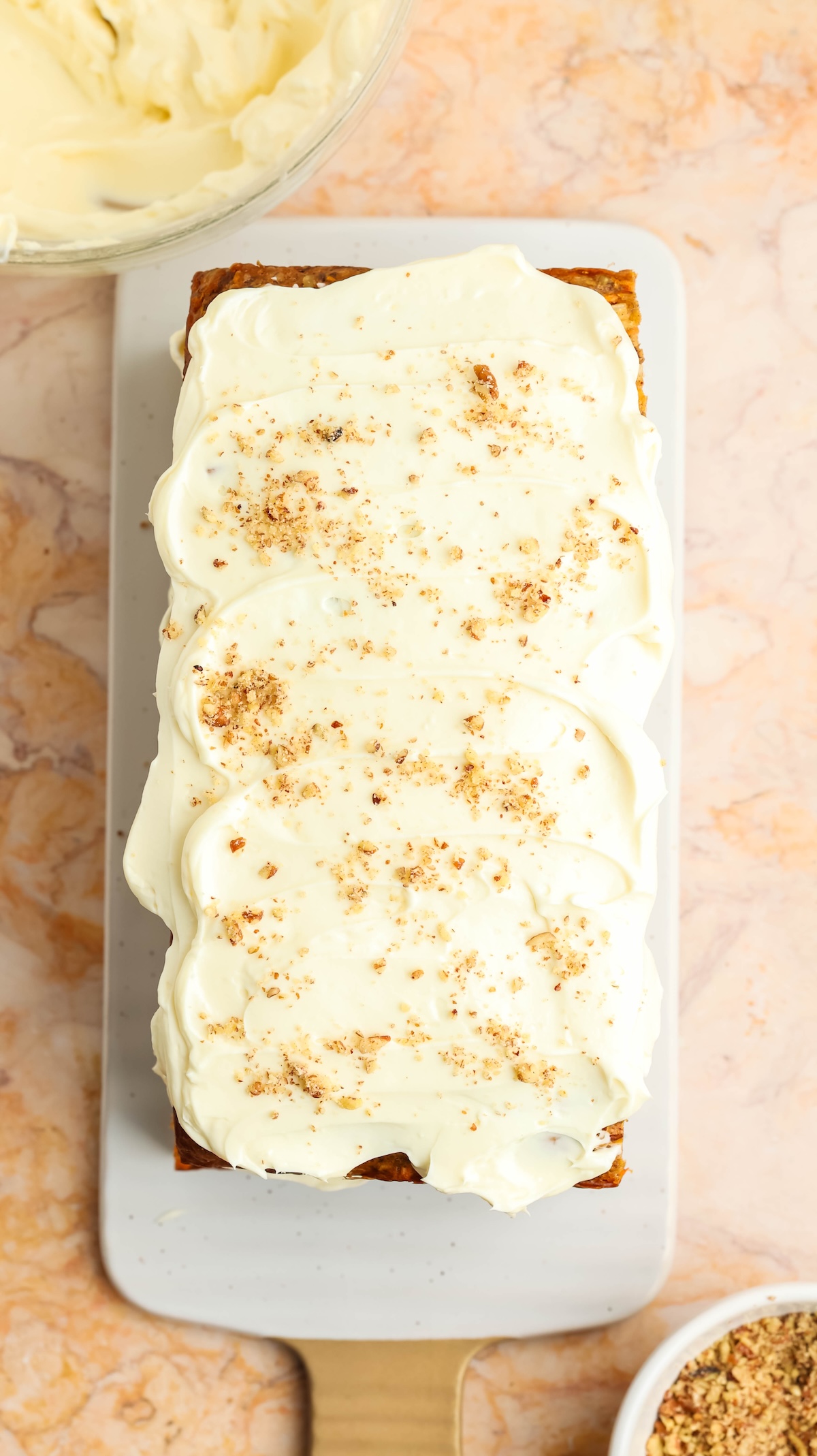 Overhead view of a rectangular carrot cake on a white serving platter, covered with thick cream cheese frosting and sprinkled with chopped nuts. A glass bowl of frosting and a bowl of nuts are visible in the background, set on a marble countertop.