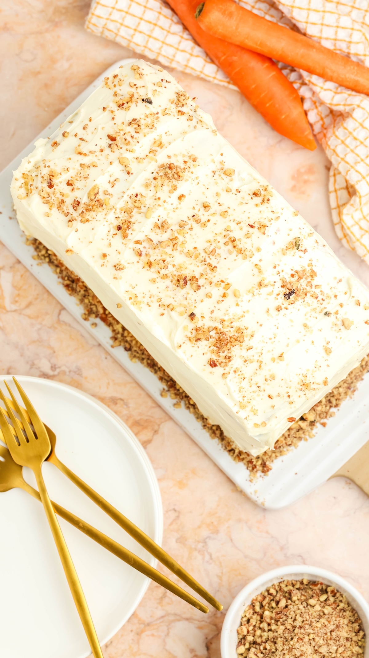Overhead view of a frosted carrot cake topped with chopped nuts on a white serving platter. The scene includes fresh carrots on a yellow and white checkered napkin, gold cutlery on stacked white plates, and a bowl of nuts, all arranged on a marble countertop.