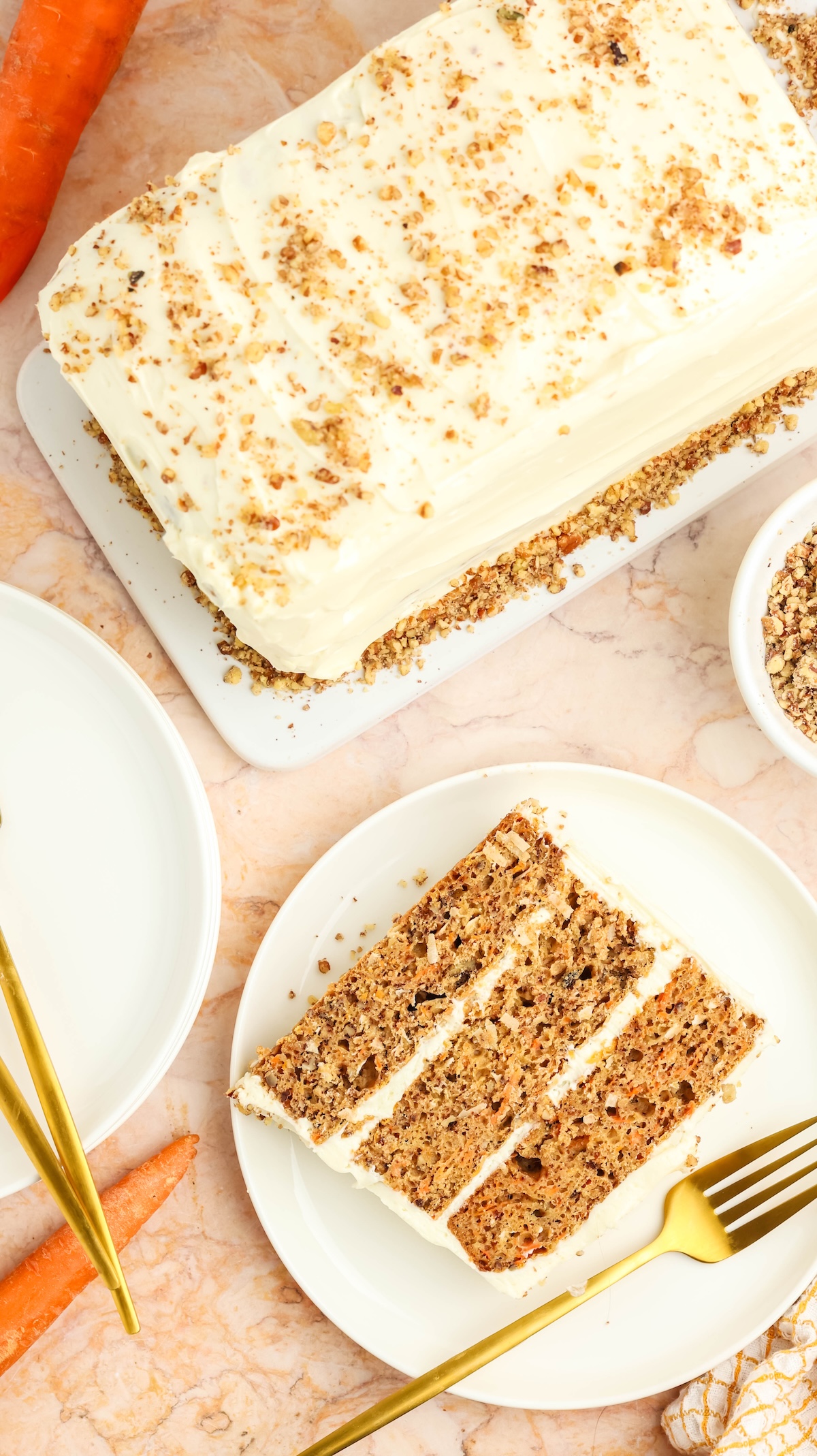 Flat lay of a frosted carrot cake topped with chopped nuts on a white platter. A slice of the cake is served on a white plate with a gold fork, showing its layered interior. Fresh carrots and a bowl of nuts are also visible, set on a marble surface.
