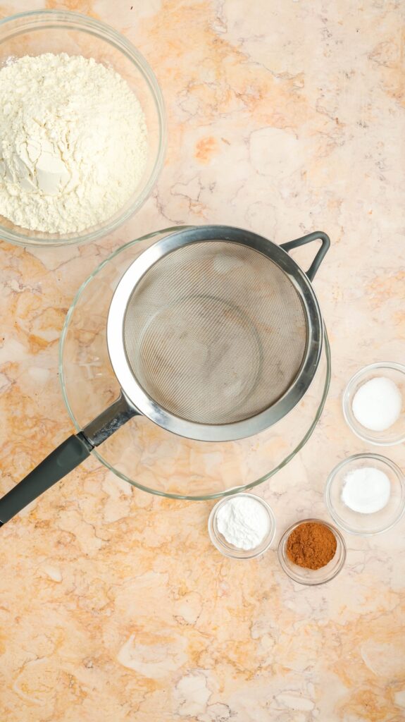 Flat lay of dry baking ingredients for carrot cake on a marble surface. Features a large bowl of flour, smaller bowls of sugar, baking powder, cinnamon, and salt, with a metal sifter positioned over an empty glass bowl.