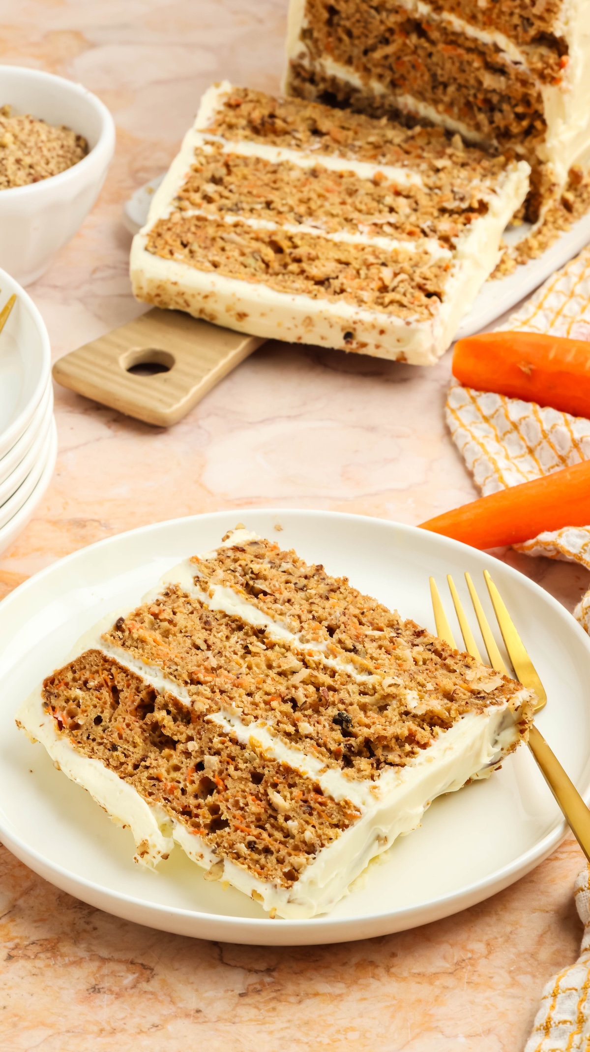 Overhead view of a slice of carrot cake with cream cheese frosting on a white plate, accompanied by a golden fork. The rest of the cake sits on a wooden cutting board in the background, with fresh carrots and a checkered kitchen towel nearby, all on a marble countertop.