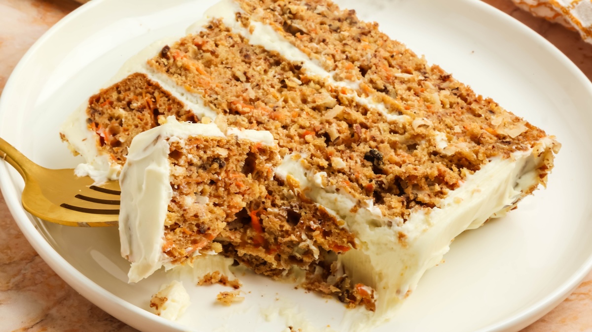 High-angle view of a slice of carrot cake with cream cheese frosting on a white oval plate. A golden fork is partially embedded in the cake, lifting a piece, with a blurred marble surface and checkered napkin in the background.