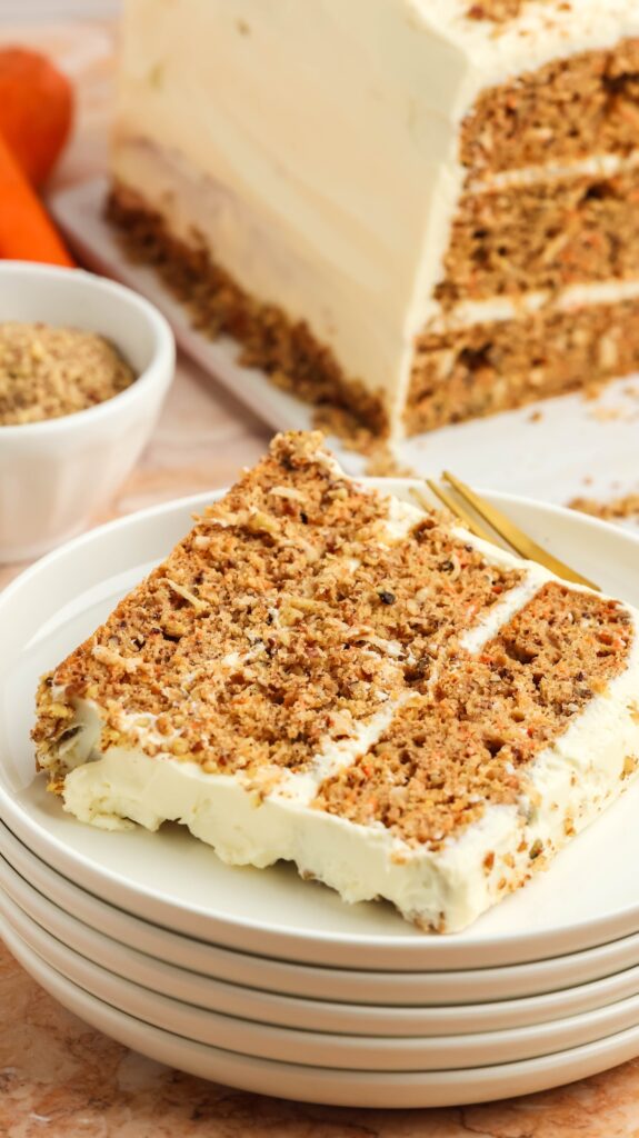 Overhead view of a slice of carrot cake with cream cheese frosting and a crumbly nut topping on a white plate. A golden fork rests beside the slice, with the whole cake and fresh carrots visible in the background, set on a marble surface.