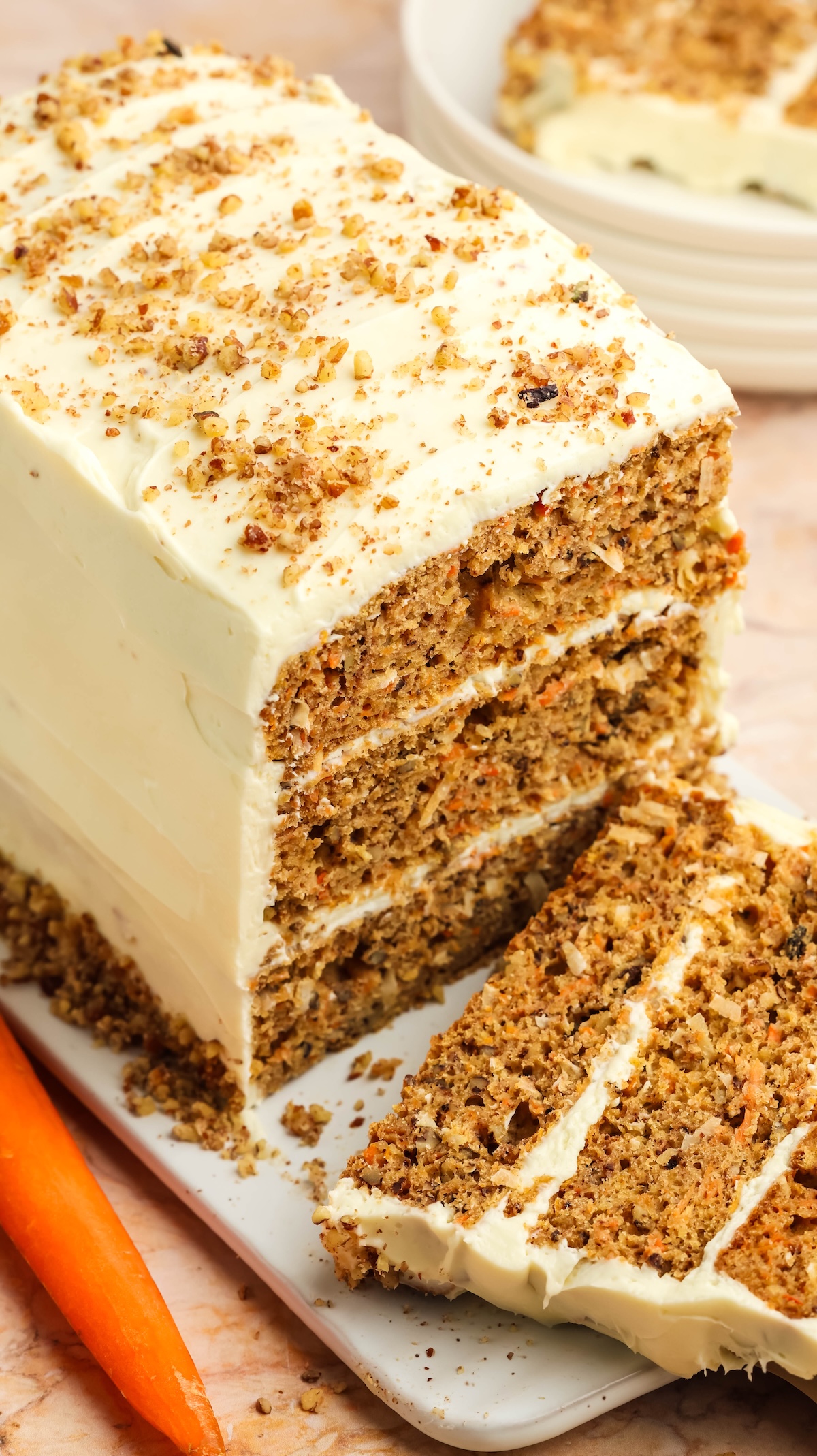 Angled view of a rectangular carrot cake with cream cheese frosting and chopped nuts on a white serving platter. A slice has been cut and placed next to the cake, revealing its moist layers, with a fresh carrot in the foreground.
