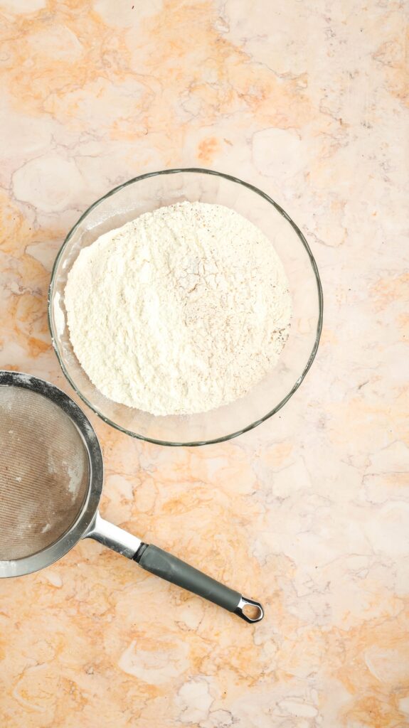 Top-down view of a glass bowl containing a mixture of flour and spices, alongside a metal sieve with a black handle, set on a light marble countertop, ready for sifting.