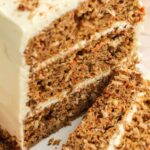 Vertical close-up of a multi-layered carrot cake with cream cheese frosting on a white serving platter. Two slices are cut and laid on their sides, showcasing the moist layers, frosting, and nut topping, with a soft-focus background.