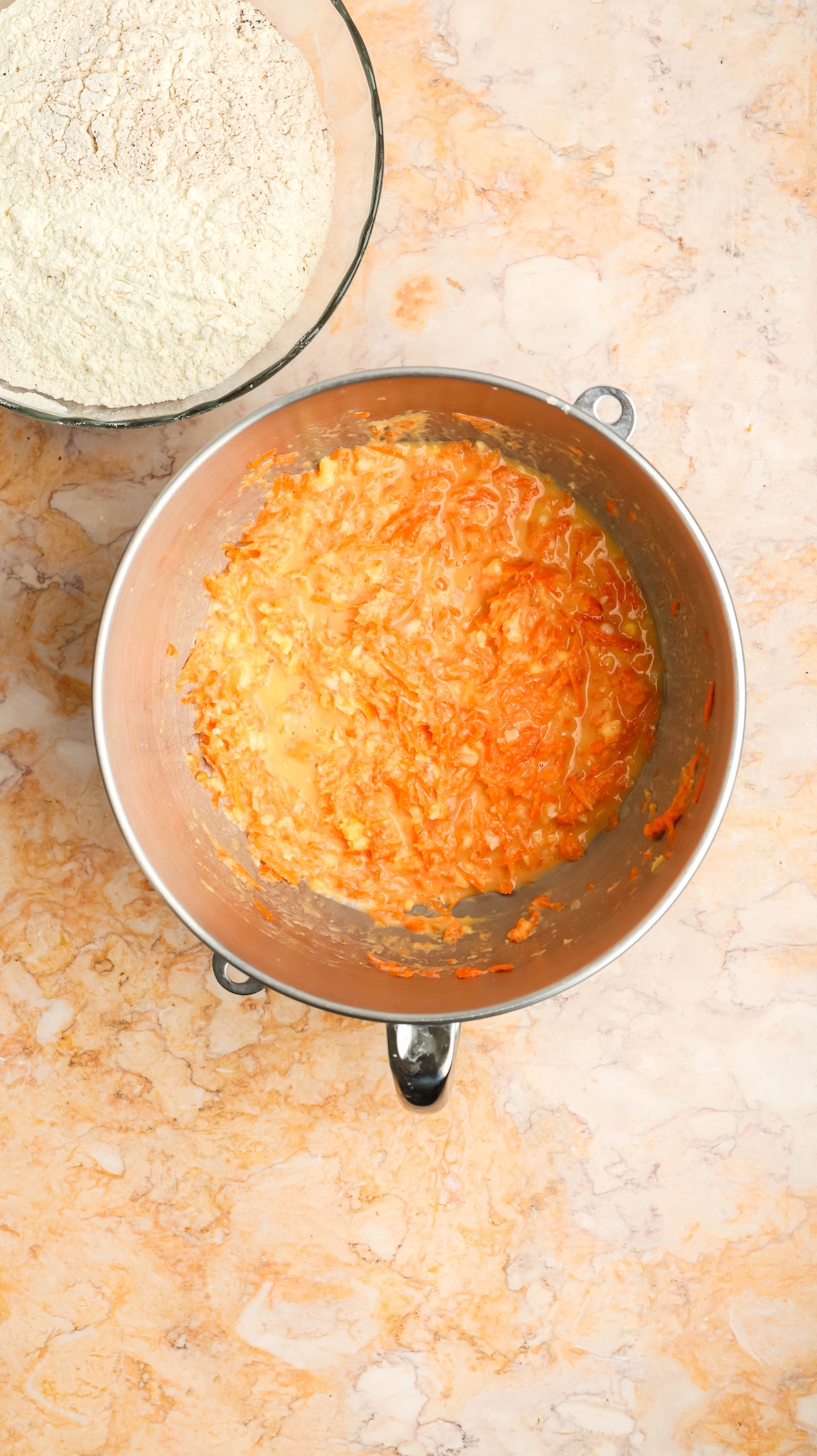 Overhead shot of two bowls on a marble surface: a glass bowl with flour and a large metallic bowl containing a vibrant orange carrot cake batter with visible shredded carrots.