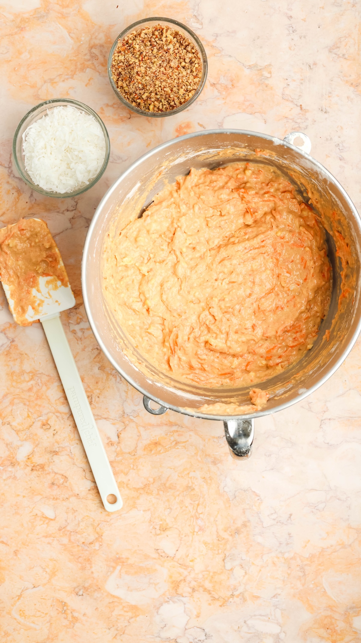 Flat lay of a large metallic mixing bowl filled with carrot cake batter, a white spatula with batter residue, and two small bowls containing shredded coconut and chopped nuts, all set on a warm-toned marble surface.