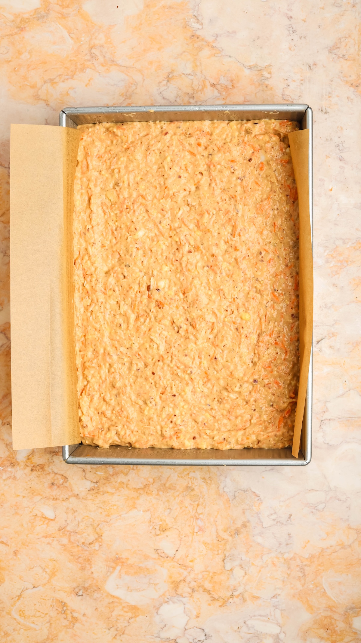 Overhead view of a rectangular baking pan lined with parchment paper and filled with thick, orange carrot cake batter. The pan is placed on a light marble surface with warm tones.