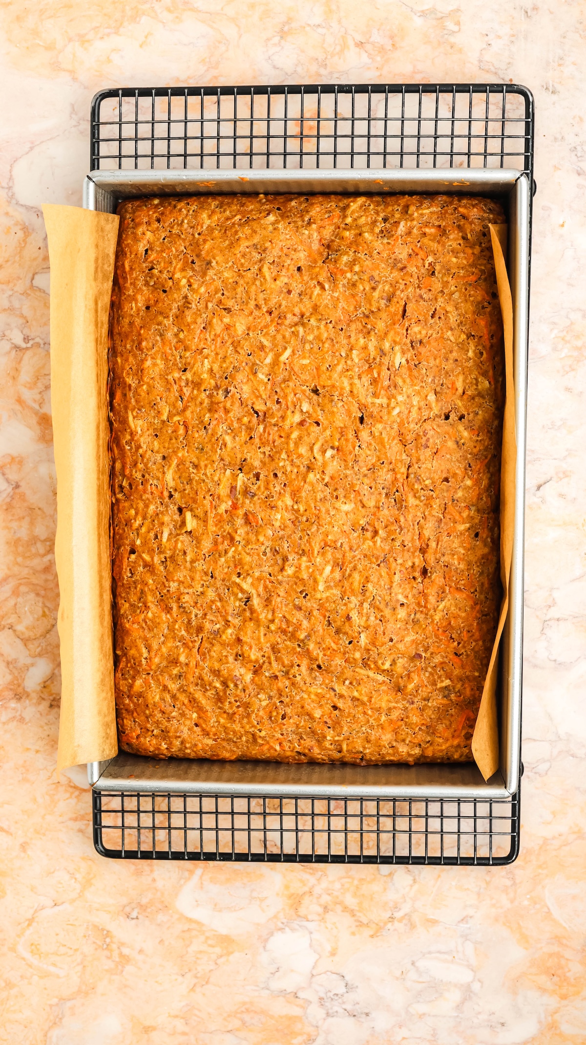 Top-down view of a freshly baked rectangular carrot cake in a silver baking pan, resting on a black wire cooling rack. The pan is lined with parchment paper, and the setup is on a light marble countertop.