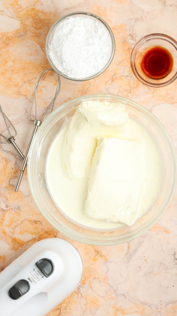 Flat lay of baking tools and ingredients for cream cheese frosting on a marble surface. Includes a large glass bowl with cream cheese and heavy cream, a smaller bowl of powdered sugar, a bowl of vanilla extract, and a white hand mixer with metal beaters.