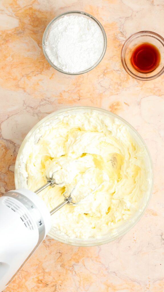 Overhead shot of a glass bowl containing pale yellow cream cheese frosting being mixed with a white hand mixer. Nearby are smaller bowls of powdered sugar and vanilla extract, all set on a light marble countertop.