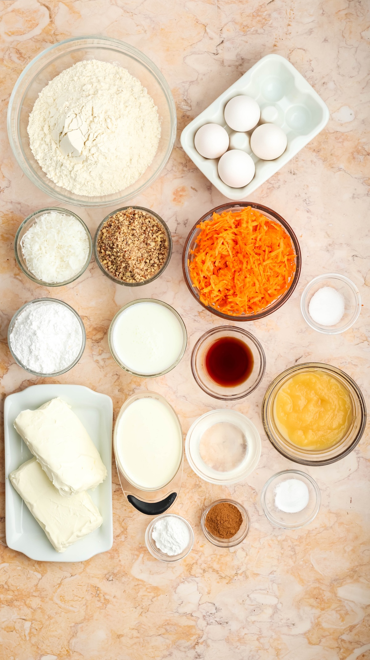 Overhead view of carrot cake ingredients arranged on a light marble surface. Includes bowls of flour, shredded carrots, coconut, chopped pecans, cream cheese, eggs, applesauce, buttermilk, and spices, all pre-measured and ready for baking.