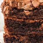 Close-up of a stack of three rich, dark chocolate brownies with crinkly tops and dense, fudgy interiors. The brownies are topped with scattered milk chocolate chunks, and the soft, light background highlights their indulgent texture.