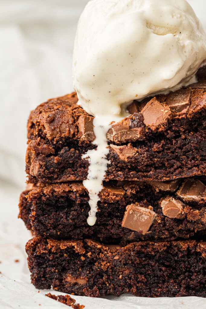Close-up of a stack of three chocolate brownies topped with a scoop of vanilla ice cream. The brownies have a rich, fudgy texture with crinkly tops and are adorned with milk chocolate chunks.
