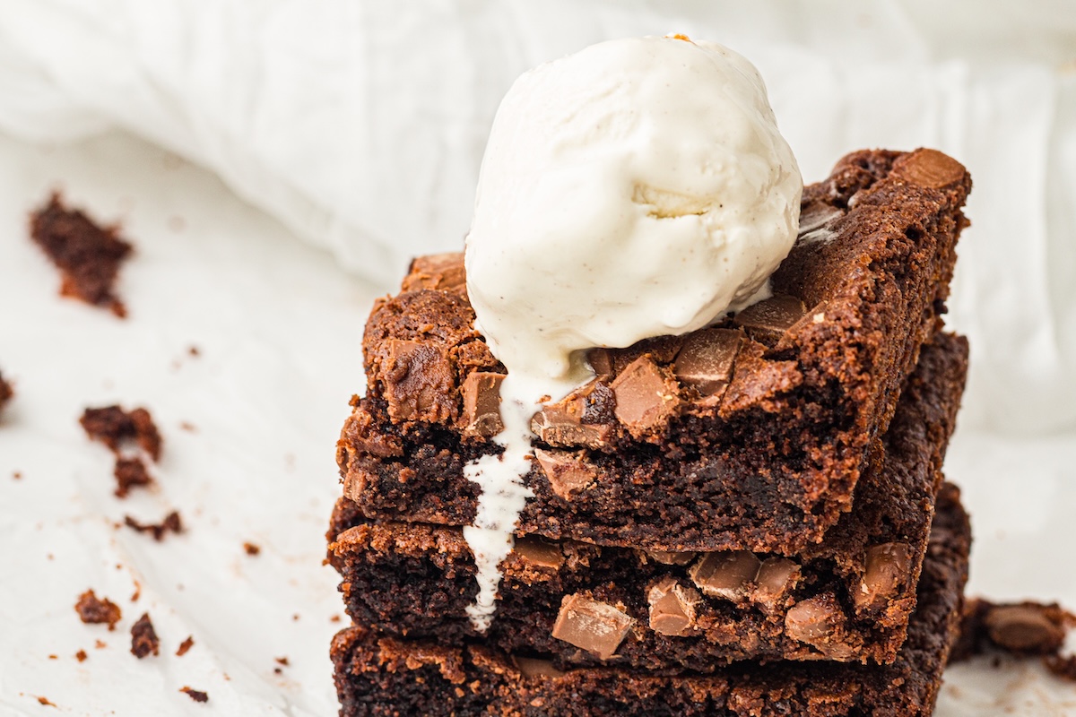 Close-up of a stack of three chocolate brownies topped with a melting scoop of vanilla ice cream. The brownies are dark brown with a fudgy texture and crinkly tops, adorned with scattered milk chocolate chunks.