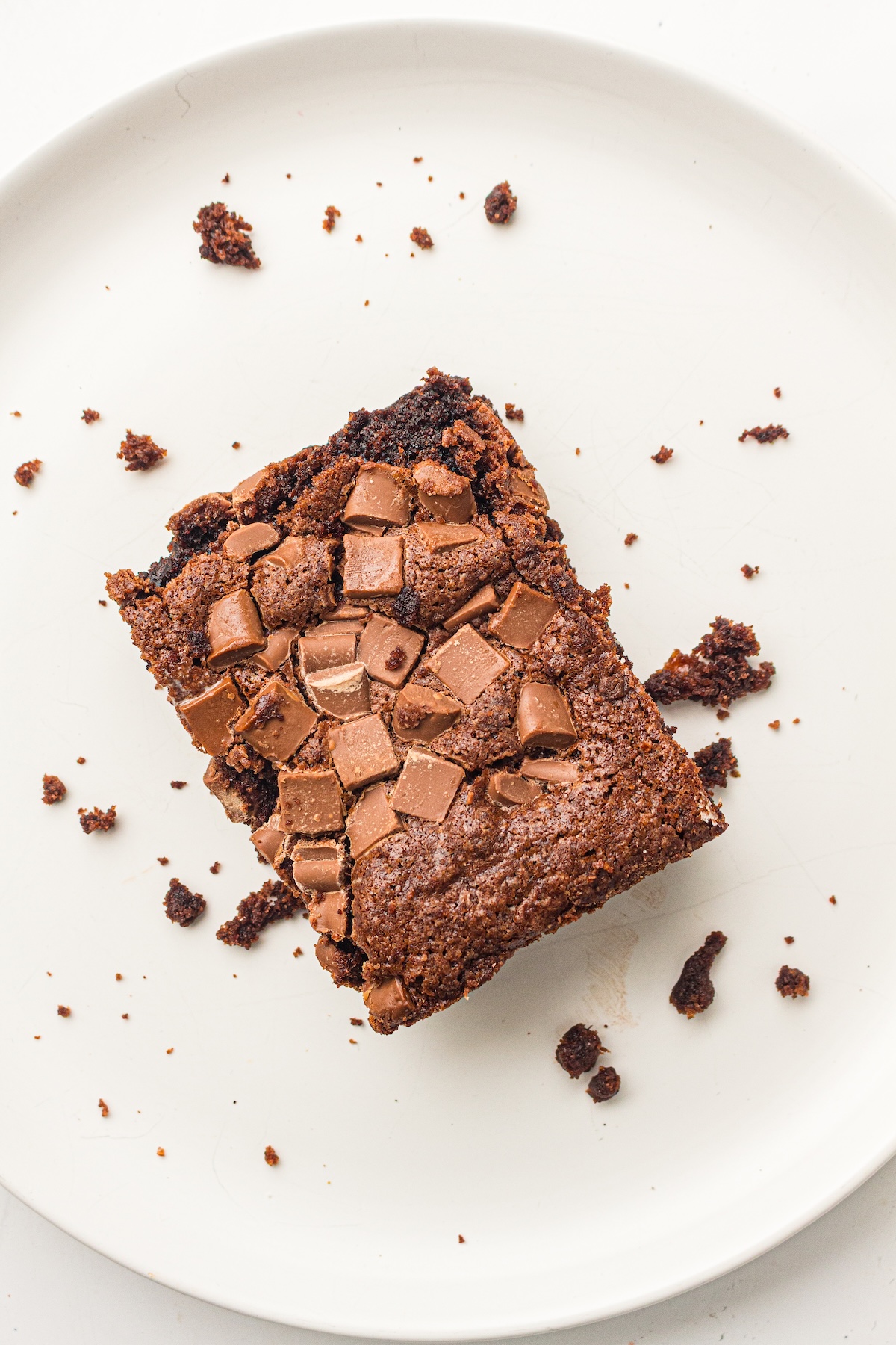 Top-down view of a single chocolate brownie square on a plain white plate. The brownie has a dense, fudgy texture with a crinkly top and is generously topped with milk chocolate chunks.