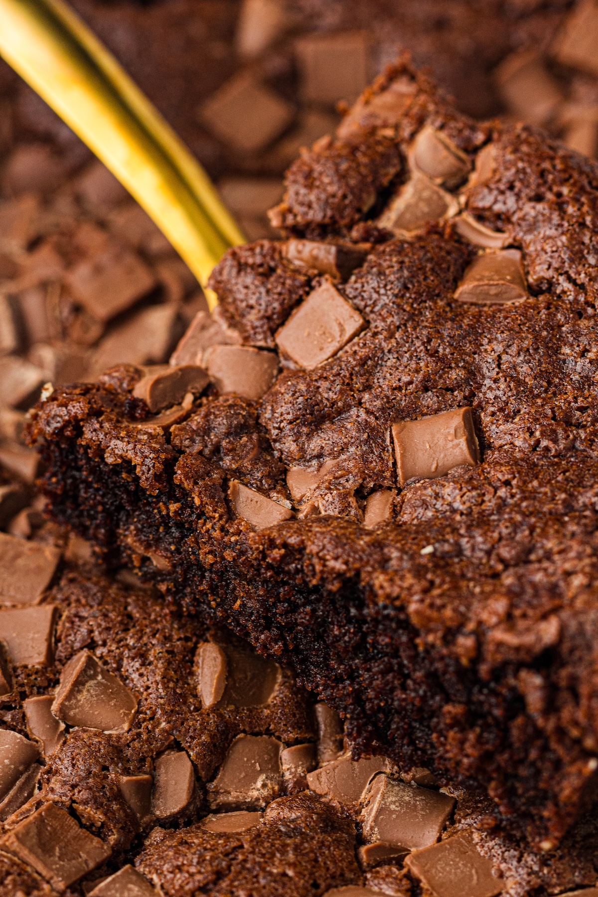 Close-up of a freshly baked chocolate brownie in a white baking dish. A rectangular piece is prominently displayed, showing its dense, moist interior. The top is crinkly and studded with milk chocolate chunks, with a golden serving utensil partially visible underneath.