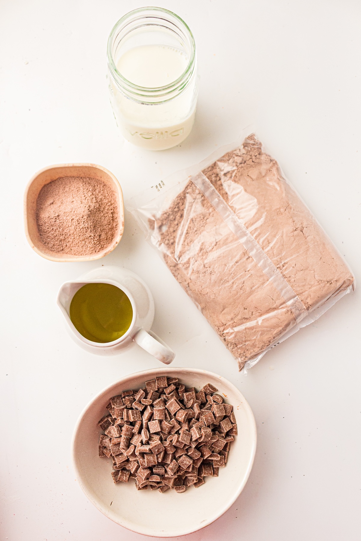 Top-down view of chocolate dessert ingredients arranged on a white surface. Includes a bowl of cocoa powder, a mason jar of milk, a bag of chocolate mix, a creamer of oil, and a bowl of chocolate chunks.