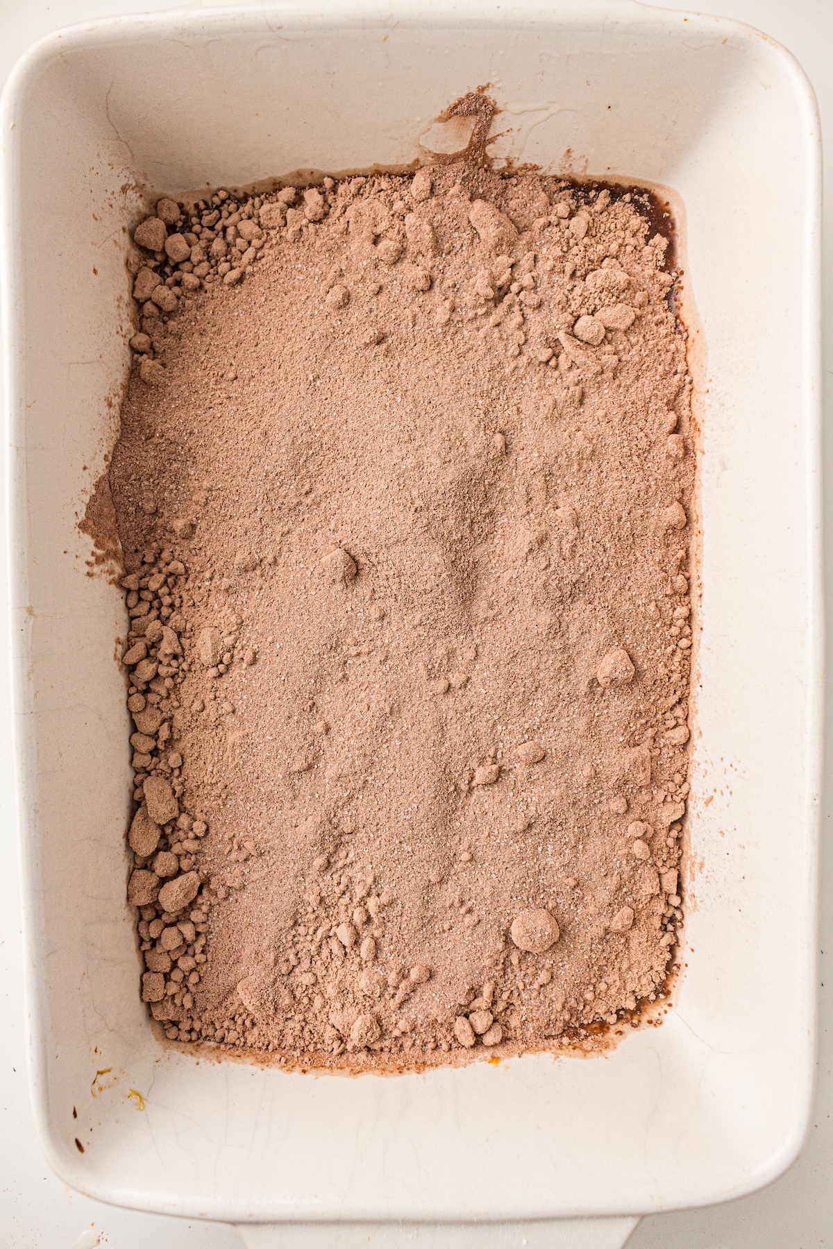 Top-down view of a white baking dish filled with a layer of light brown dry cake mix. The mix is unevenly spread, with clumps and fine powder visible, suggesting the initial stage of a dessert preparation.