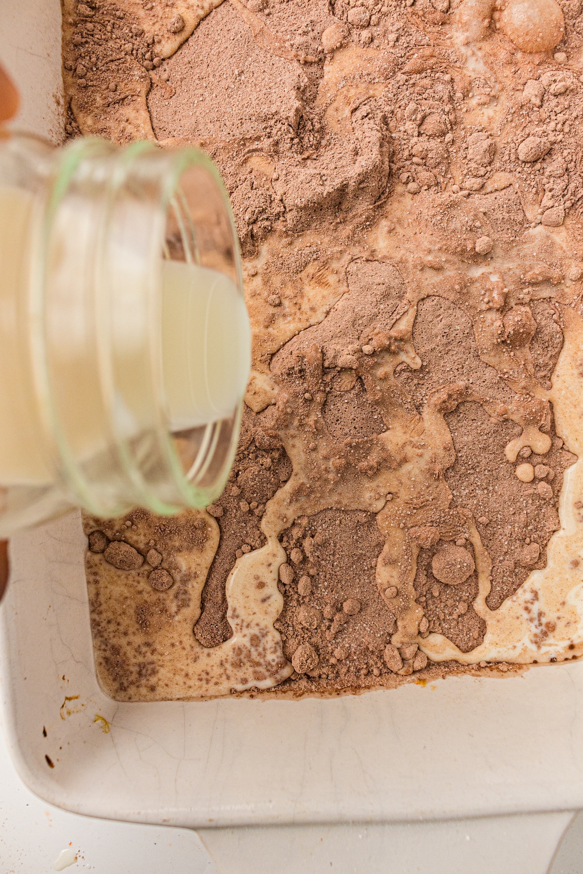 Top-down view of a light-colored liquid being poured from a glass bottle into a white baking dish filled with dry chocolate cake mix. The liquid begins to mix with the powder, creating wet patches and a darker brown paste.