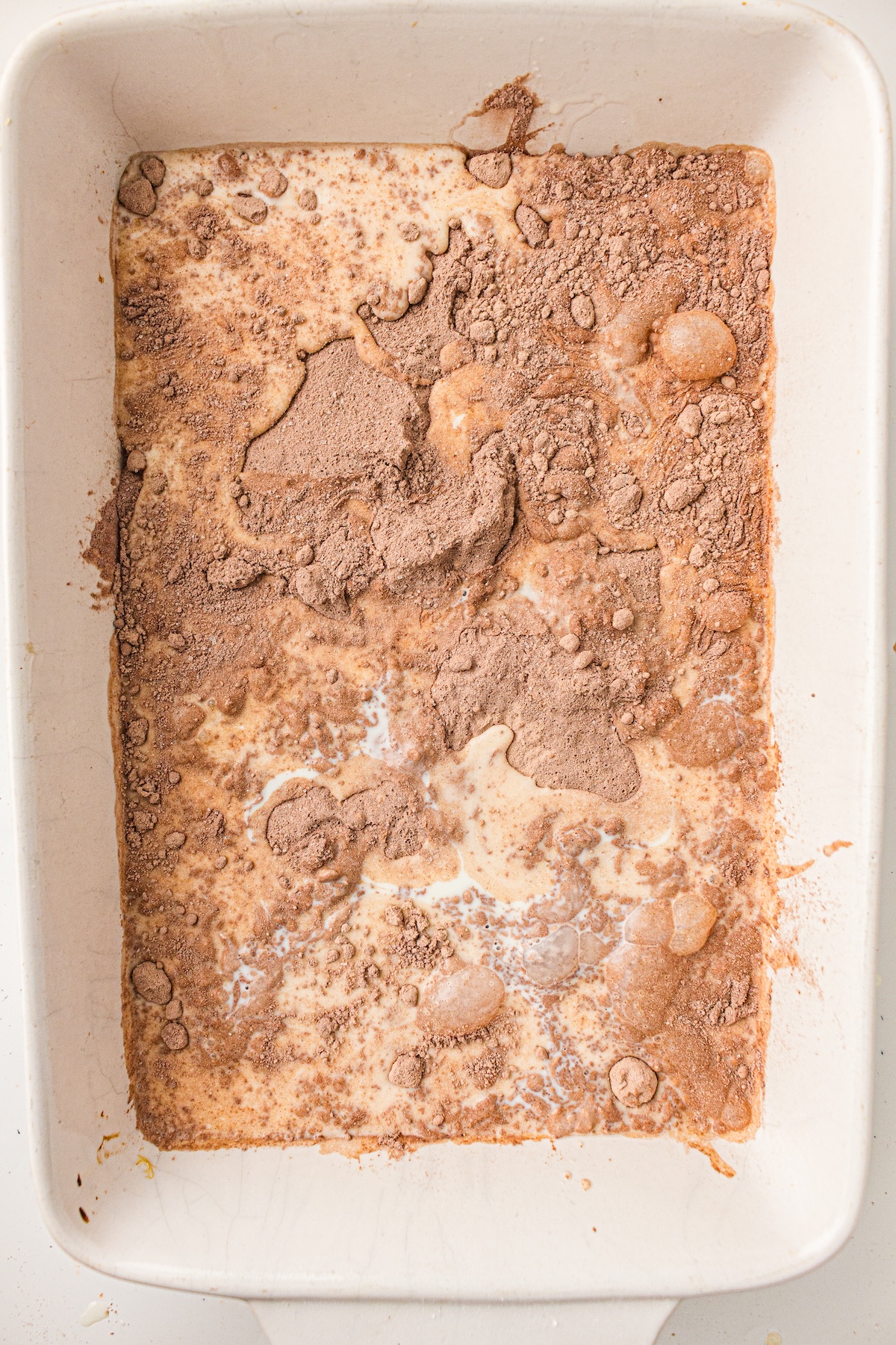 Top-down view of a creamy light brown liquid mixture in a white baking dish, partially covered with clumps and specks of dark brown cocoa powder. The uneven distribution and visible bubbles suggest an intermediate stage of dessert preparation.
