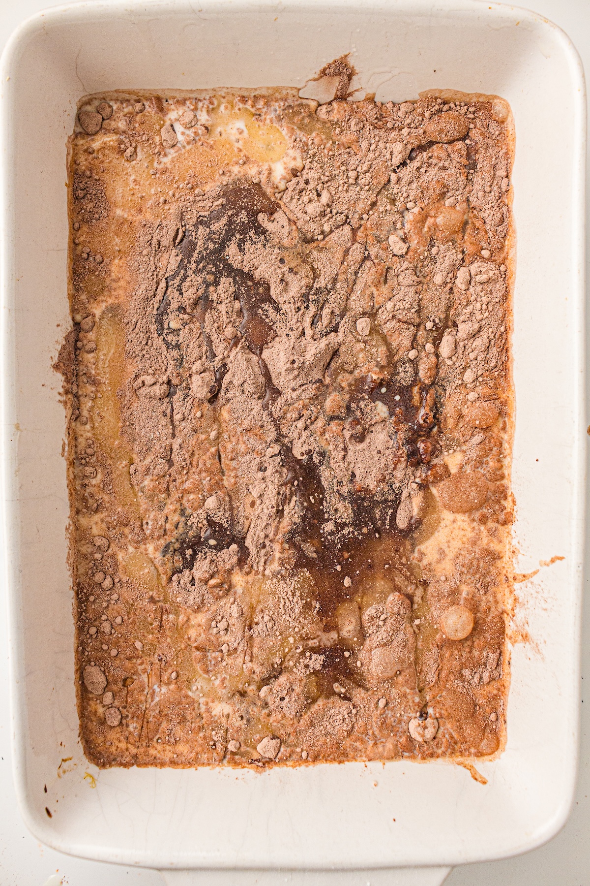 Top-down view of a white rectangular baking dish containing a mixture of light brown powder, darker wet patches, and some yellowish liquid. The uneven distribution and clumps suggest an early stage of dessert preparation, likely before mixing.