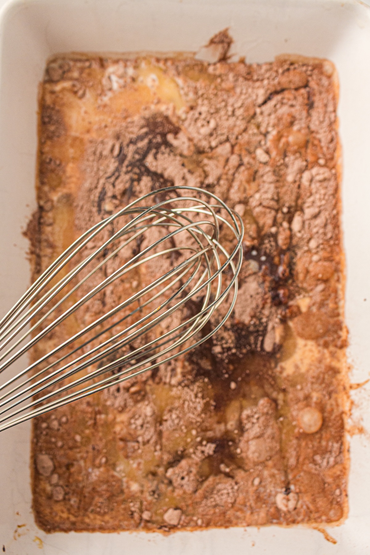 Close-up of a white rectangular baking dish with a brown powdery mixture and yellowish liquid patches. A silver whisk rests in the dish, indicating the mixing stage of a dessert preparation. The uneven texture highlights the combination of dry and wet ingredients.